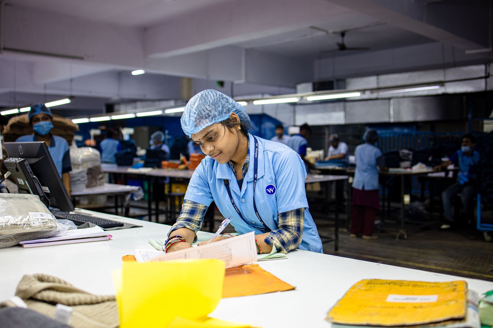 Indian factory worker in uniform and hairnet recording quality checks, representing food-hygiene and QC documentation on a paper converting line.