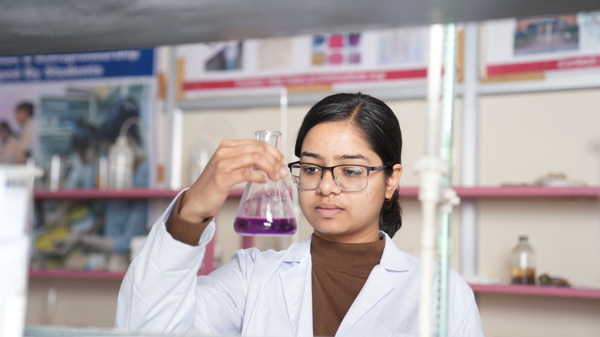 Indian chemist in a white lab coat holding up a conical flask of purple solution in a college chemistry laboratory