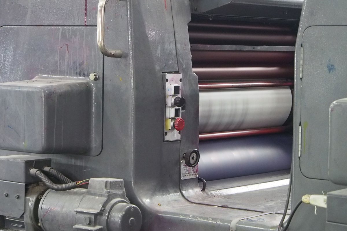 Close view of the inked roller train inside an offset printing press, with a guarded housing and a red stop button on the side frame