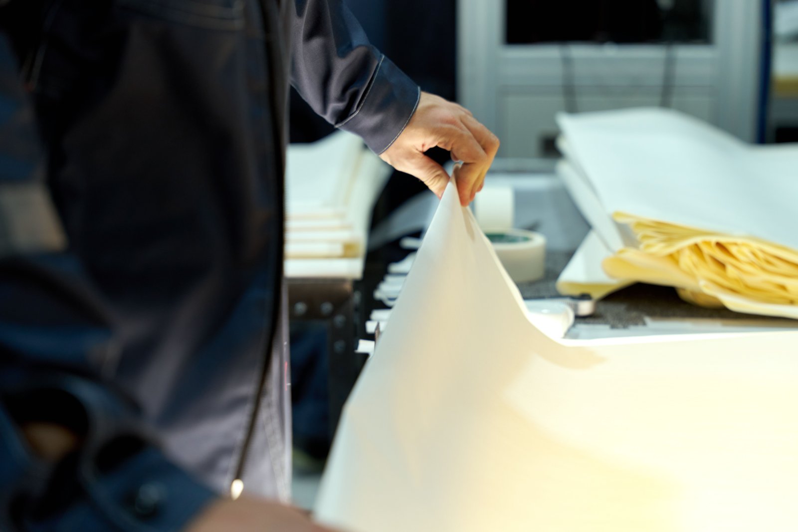 Worker in dark overalls lifting the corner of a large white sheet of paper on an inspection table