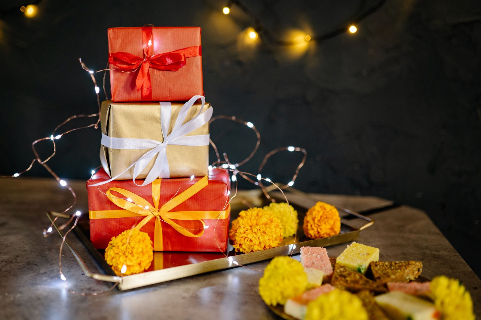 Gift boxes wrapped in red and gold paper on a brass tray with marigold flowers, fairy lights and Indian sweets for Diwali