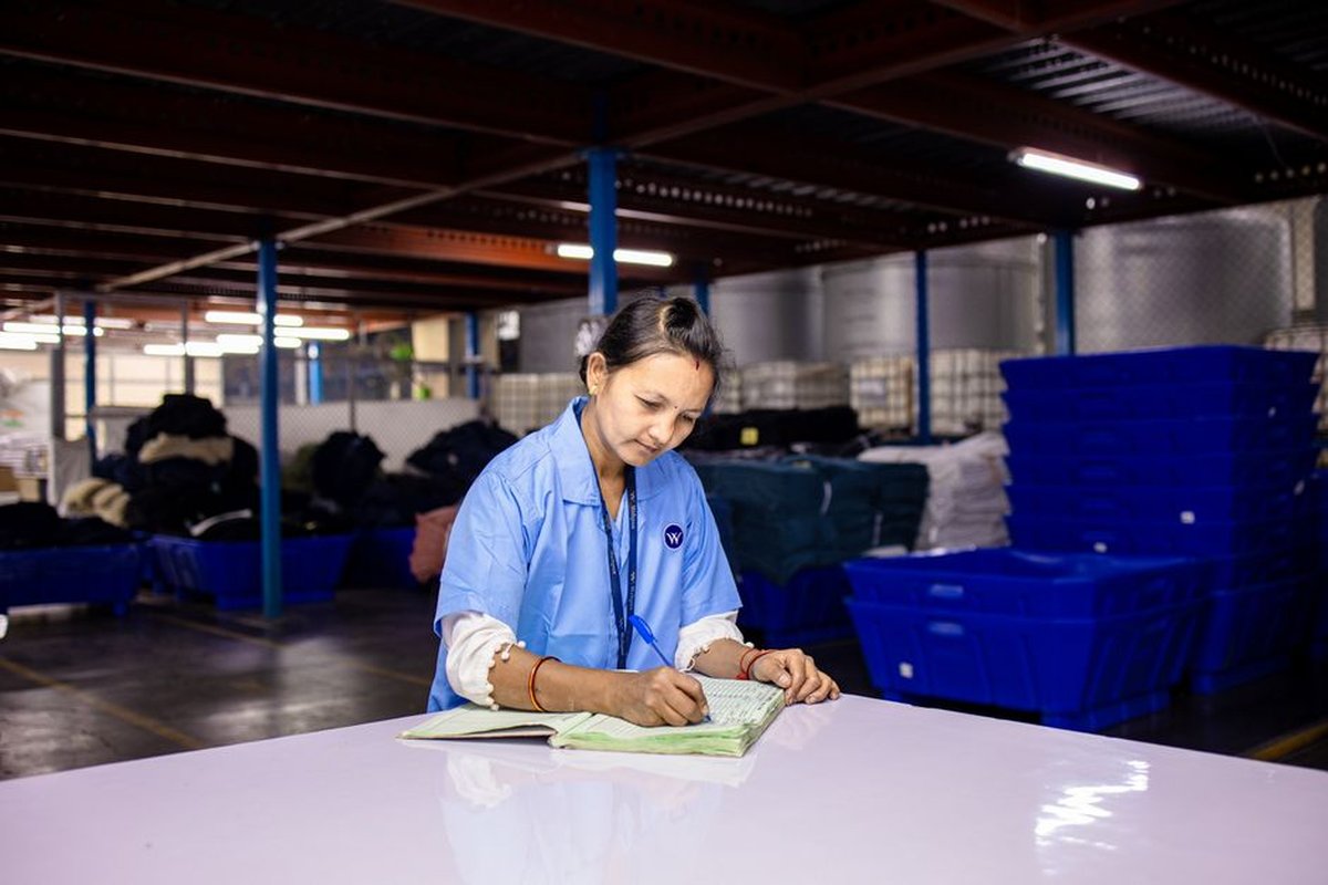 A worker records production data by hand in a logbook at a warehouse workstation in India