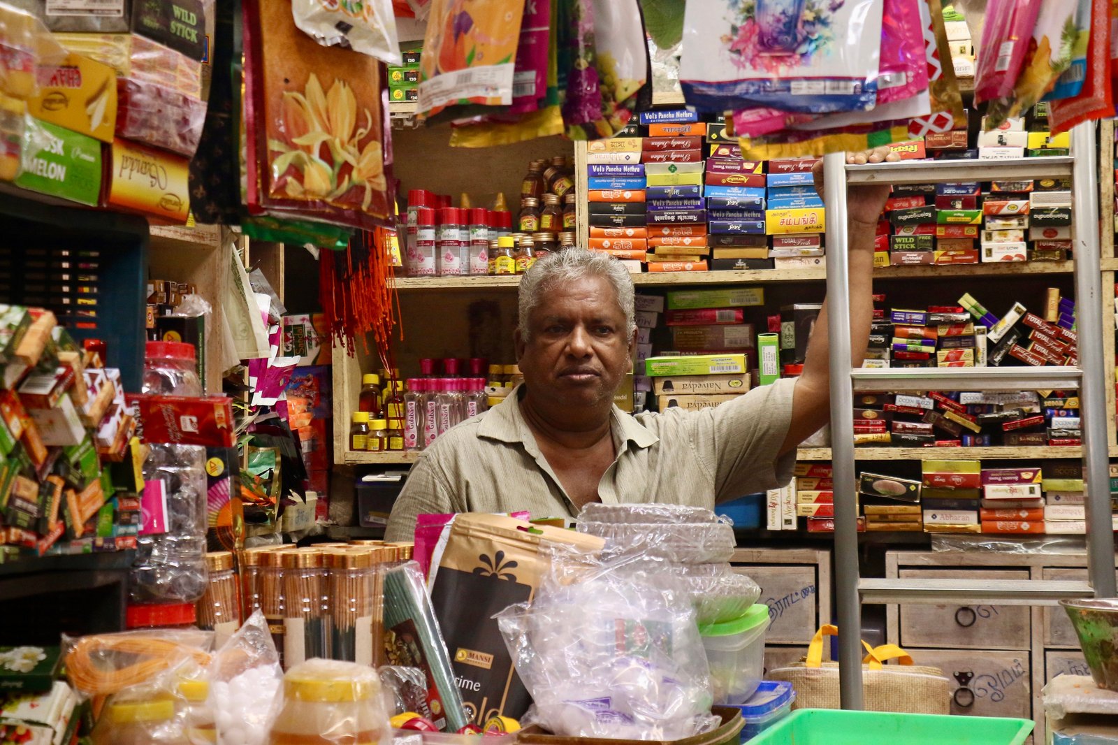 A traditional Indian kirana store owner behind a counter stocked with packaged foods, festive decorations and carry bags