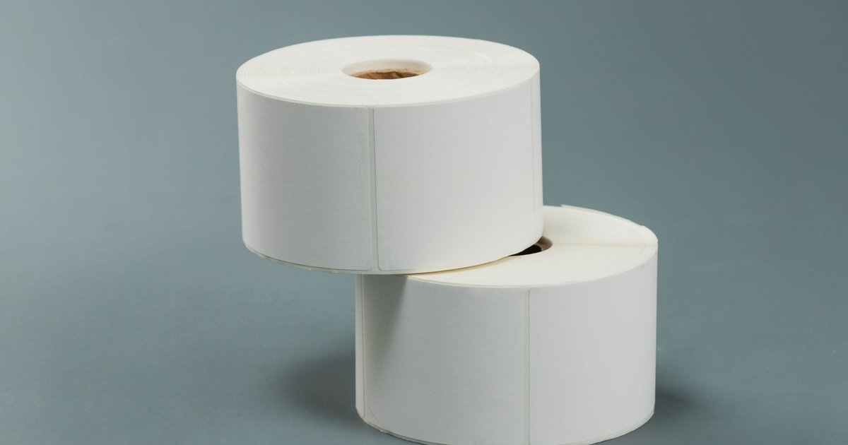 Two rolls of blank white self-adhesive labels on a release liner, stacked on a grey surface