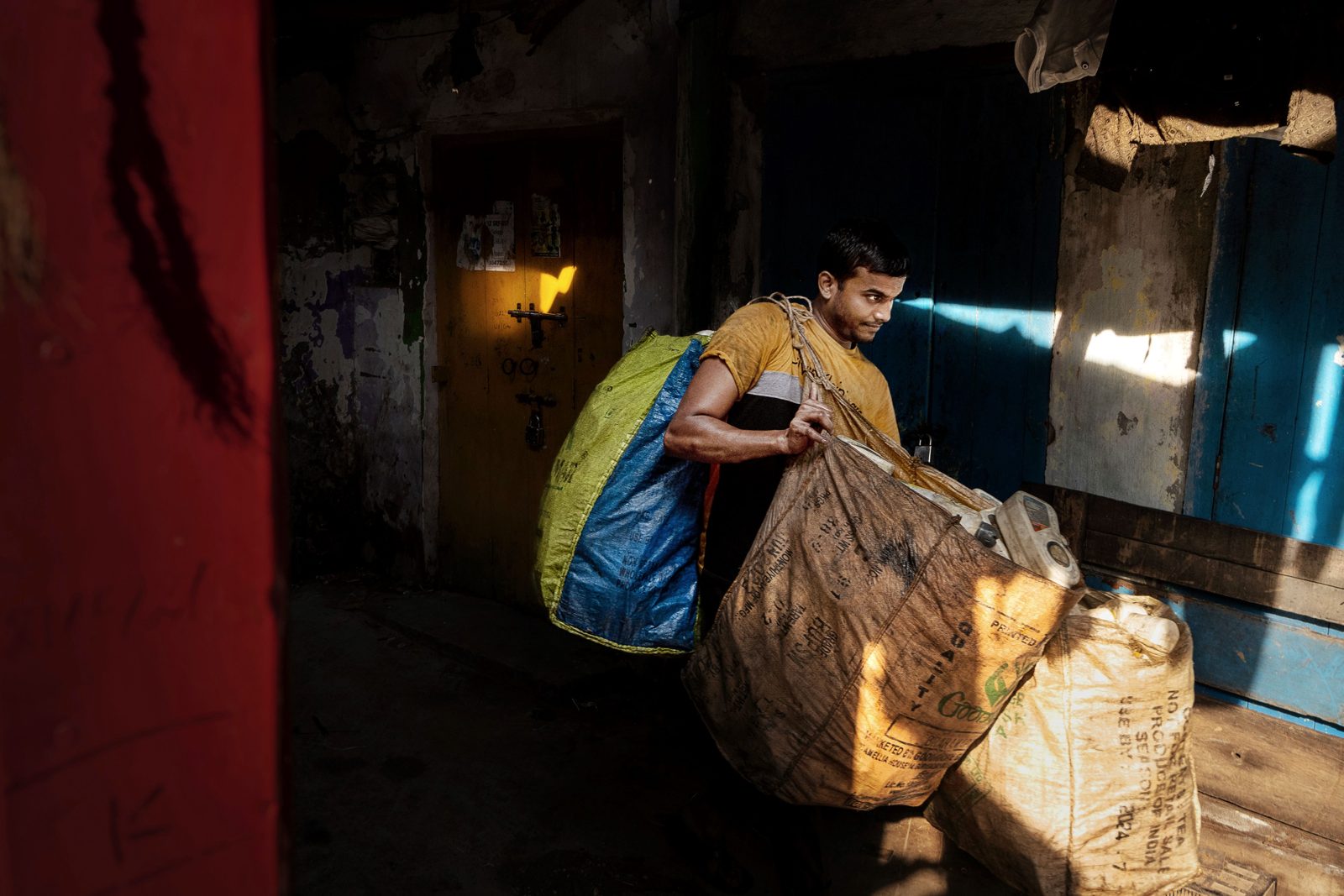 Young man in India carrying large woven sacks of collected scrap through a narrow doorway